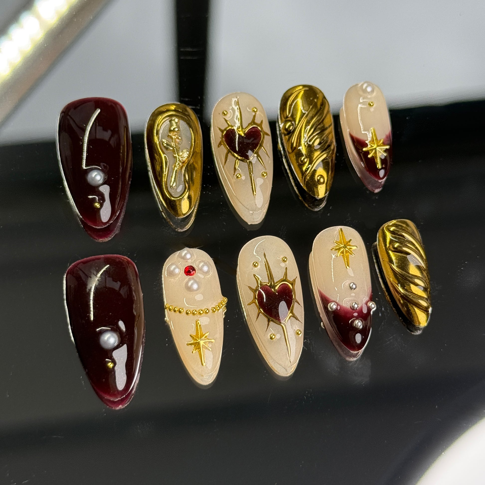 Burgundy and gold nail set with heart motifs, pearls, and star embellishments, displayed on a reflective surface.